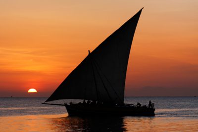 En traditionell segelbåt heter dhow på Zanzibar. Silhuetten av en dhow utmärker sig mot en brandgul solnedgång.
