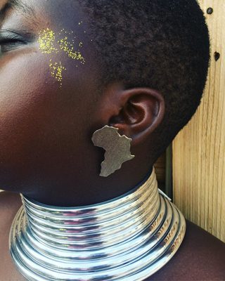 Julklappstips 2021 för den som älskar smycken och Afrika, ett örhänge med Afrikas karta utskuret i mässing. Örhänget syns på en svart kvinna med glitter i ansiktet, som delvis är bortvänt, och ett tjockt silvrigt halsband.