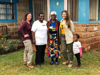 Två vita kvinnor står uppställda tillsammans med en kenyansk familj framför ett hus. Upplev Kenya som en äkta kenyan och bo hos en värdfamilj under ditt besök
