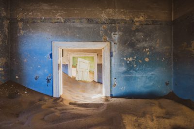 Vi ser ett rum med flagnande blå färg. I mitten finns en dörröppning och det syns flera dörröppningar inuti den. Det är sand halvvägs uppför rummet.