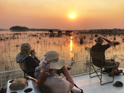 Några personer med safariklädsel sitter på en båt, solnedgången speglar sig i vattnet och det syns tre silhuetter av elefanter. Personerna befinner sig på Zambezifloden, på resa till Zambia.
