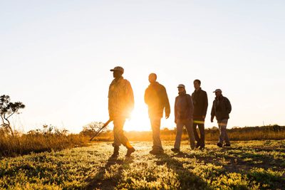 En guide med gevär går framför fyra turister som är på safari till fots under sin resa till Zambia. Guiden och turisterna syns som siluetter framför en soluppgång