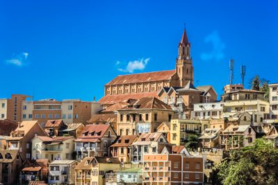 Antananarivo, i folkmun kallad "Tana", har många byggnader som är byggda på stans sluttningar. På bilden finns ett flertal bostadshus med en kyrka i bakgrunden, och en blå himmel.