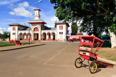 En pousse pousse, röd ricksha eller cykeltaxi, framför Antsirabes gamla tågstation. Tågstationen är en vacker byggnad i gammal kolonial stil.
