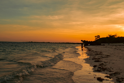 En turist vandrar längs stranden i Watamu i solnedgången, det är orangea toner från solen som reflekteras i havet.