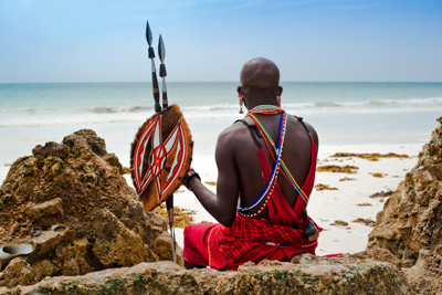 en maasai man i krigskläder sitter på Diani Beach, i Kenyas södra kust, med ryggen mot kameran och tittar ut över havet