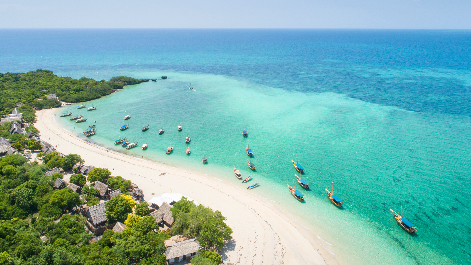 Zanzibar från ovan - en lång, vit strand snirklar sig i mitten av bilden, till vänster följer boenden och växtlighet och till höger i bild ett azurblått hav med en mängd små fiskebåtar. Bilden är tagen snett från ovan.