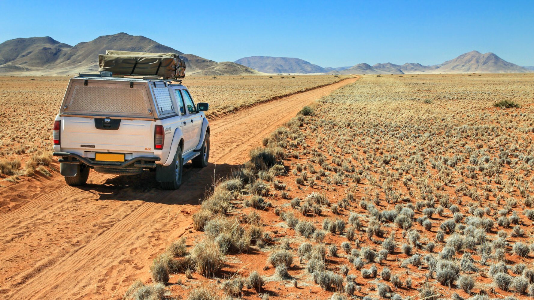En bil med fyrhjulsdrift och packning på taket kör igenom ett ökenlandskap. Grusvägen och sanden är röd, himlen är blå.