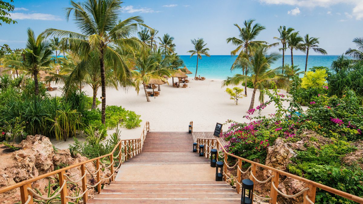 En trappa leder till en paradisstrand på Zanzibar, stranden Kendwa i norr. Det finns palmer  som skuggar solstolarna och i bakgrunden syns ett turkost hav och en ljusblå och molnfri himme.