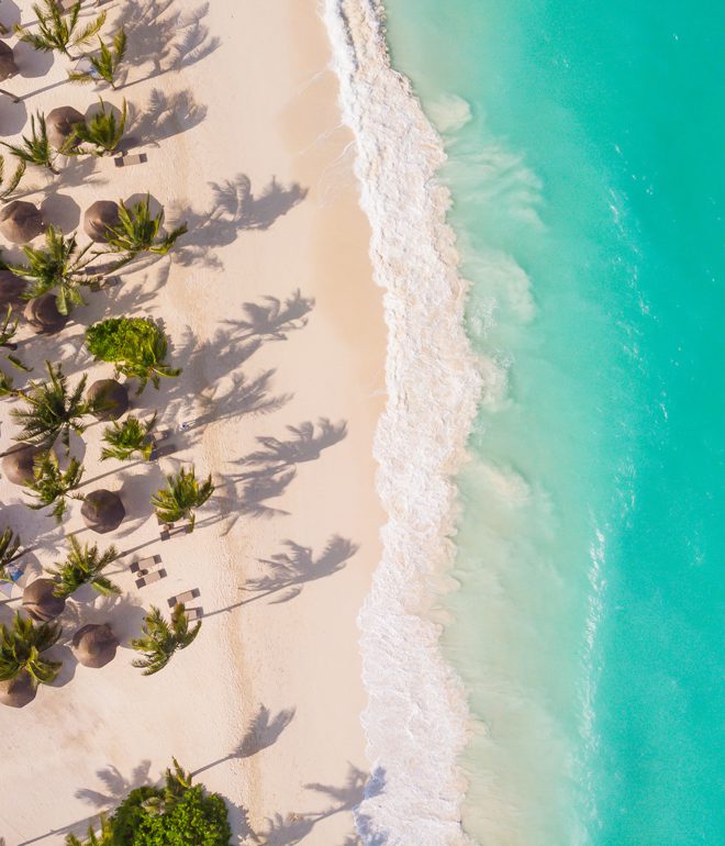 En bild på Zanzibarstranden Kendwa tagen från ovan. Till höger det turkosa havet med skummande vågor. I mitten den vita stranden och till vänster palmer och små halmtak som skyddar solstolar från solens strålar.