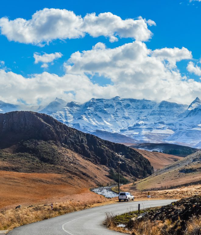 En bil kör igenom ett höstlikt landskap mot Drakensberg i KwaZulu-Natal i Sydafrika. Himlen är kornblå med tussiga vita moln och bergen är snötäckta och reser sig i bakgrunden.