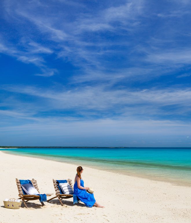 En kvinna i blå klänning sitter på en strand och tittar ut mot ett stillsamt turkost hav. Bilden är tagen på en resa till Moçambique.