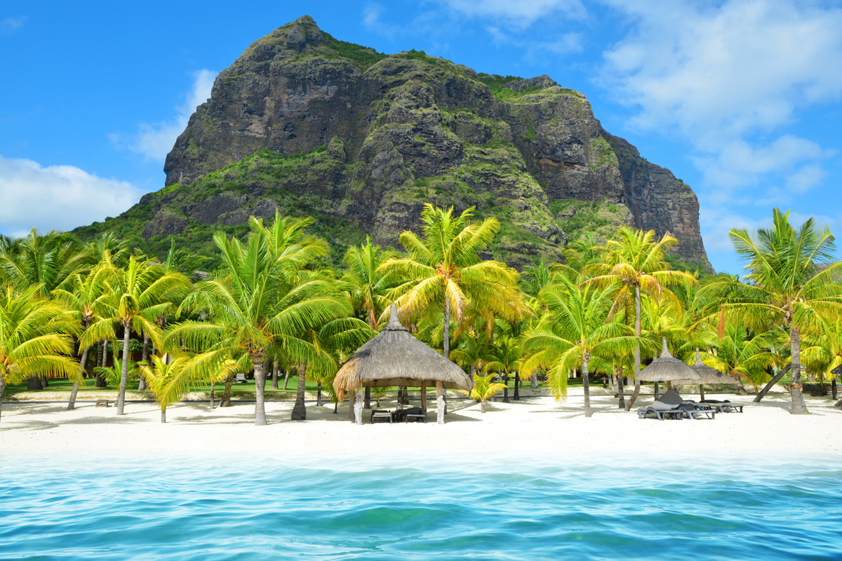 Mauritius berg Le Morne är ett UNESCO världsarv och skyddar den vackra stranden nedanför. På bilden syns det azurblå havet i förgrunden, en vit strand med några hyddor och palmer och i bakgrunden berget.